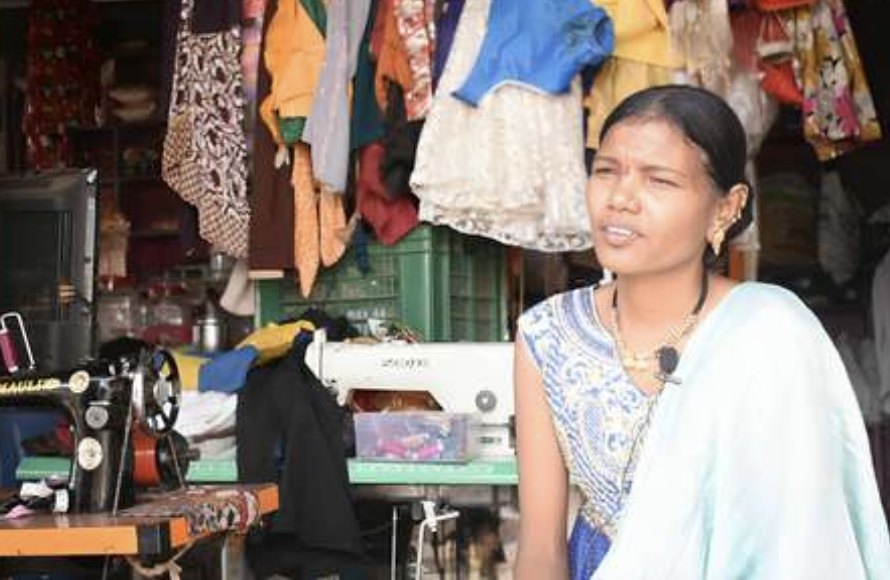 A founder in her tailoring shop, finished garments hanging behind her